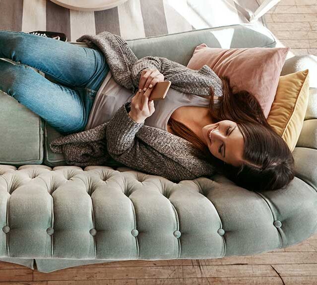 A woman laying on the couch looking at her phone with her dog laying at her feet